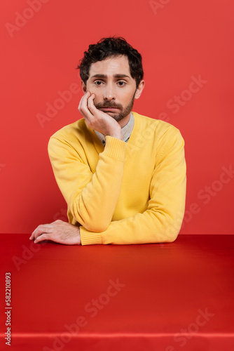 frustrated man in yellow long sleeve jumper sitting at desk on red coral background. © LIGHTFIELD STUDIOS
