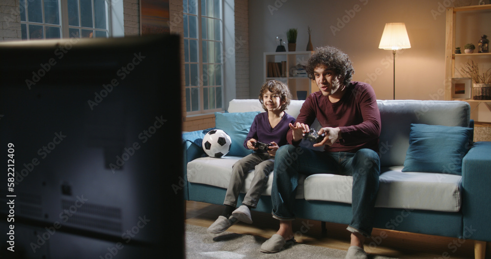 Cute authentic asian brothers with curled hair enjoying their together time, playing football video games in front of tv, parent and son relaxing with their hobby 