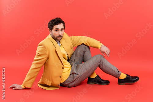 Fashionable model in yellow vest and jacket sitting on coral red background.