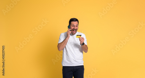 Handsome man screaming ecstatically for receiving reward while talking over smart phone and looking at credit card on yellow background