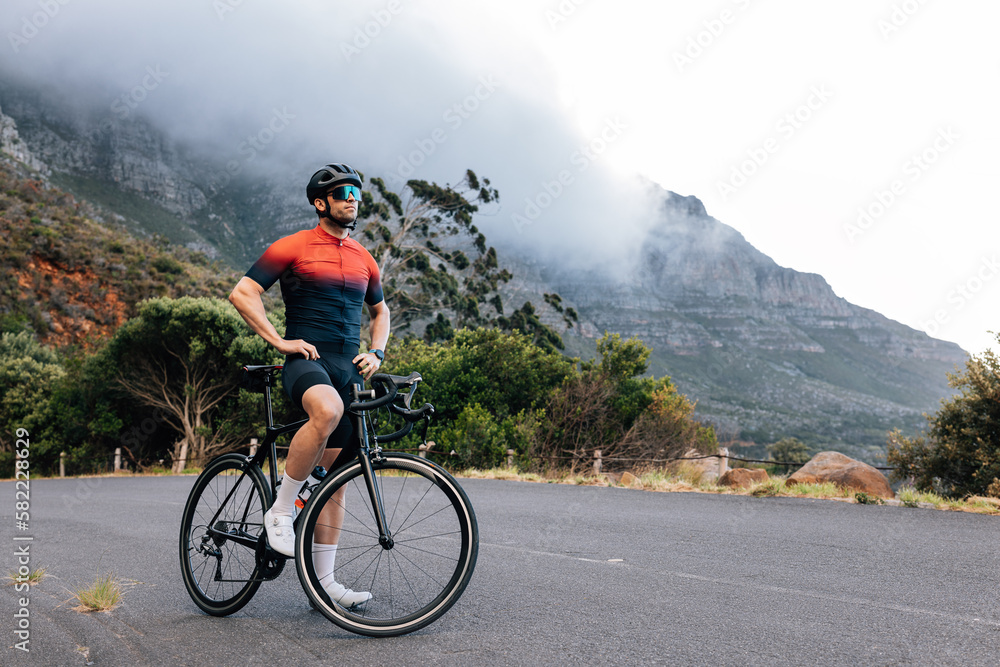 Young male cyclist relaxing after training with hands on a hips. Professional athlete with road bike enjoying the view while standing on a road in wild terrain.
