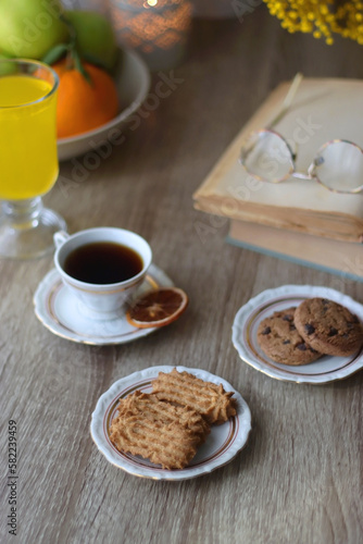 Cup of tea, plates with cookies, glass of orange juice, books, reading glasses, bowl of fruit and candles on the table. Selective focus.