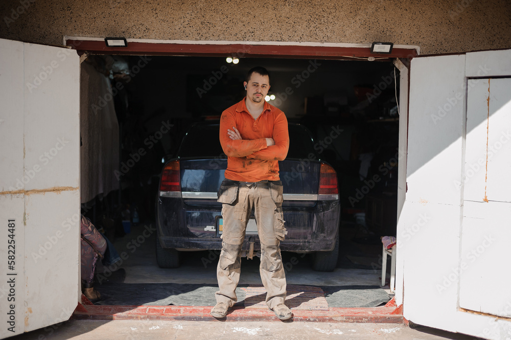 An auto mechanic stands near the garage in front of the car. A man stands near the garage in dirty work clothes An auto mechanic stands near the garage in front of the car. A man stands near the garage in dirty work clothes