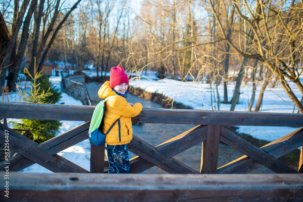 a smiling school child with a backpack in bright clothes stands on a bridge over the river, on a sunny snowy spring day
