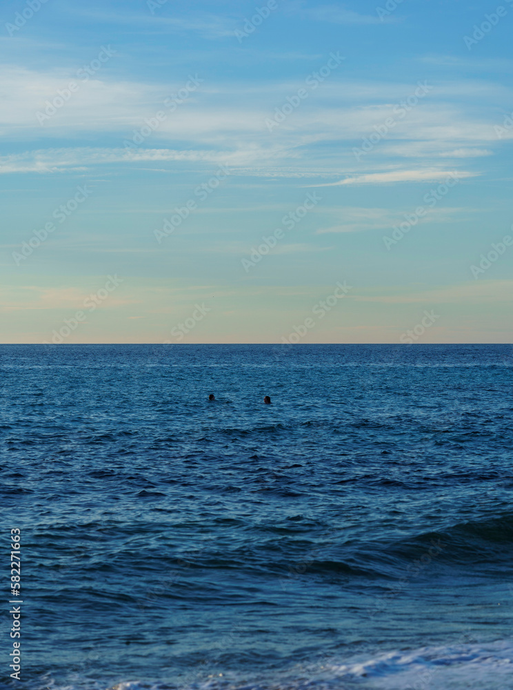 Ocean with gentle waves and two heads floating in the water, divided by clear blue sky
