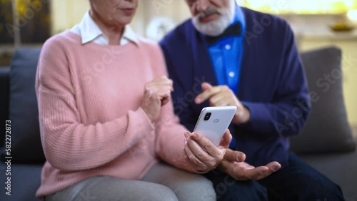 Elderly senior man and woman sitting at home on sofa grandmother holding phone showing grandfather photos scrolling news feed laughing satisfied happy 