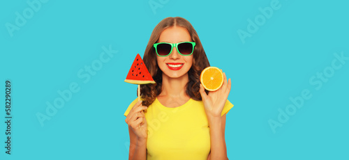 Summer portrait of happy smiling young woman with orange fruit and lollipop or ice cream shaped slice of watermelon wearing sunglasses on blue background
