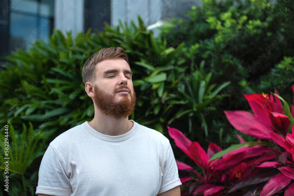 Portrait of happy young bearded man with beard breathing deep, inhale fresh air on natural background, green wall with leaves, flower in tropical country in garden. Eco, ecological concept. Portrait of happy young bearded man with beard breathing deep, inhale fresh air on natural background, green wall with leaves, flower in tropical country in garden. Eco, ecological concept.