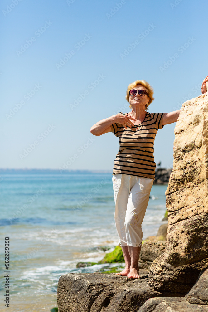 Elderly attractive smiling woman standing on a rock on the seashore waving her hand happily and enjoying life