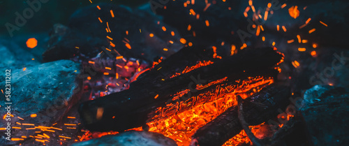 Vivid smoldered firewoods burned in fire closeup. Atmospheric background with orange flame of campfire. Wonderful full frame image of bonfire with glowing embers in air. Warm logs  bright sparks bokeh