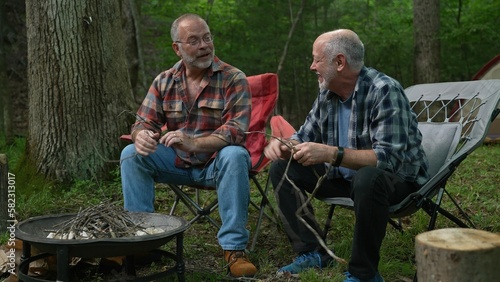 Two gay men building a campfire in a forest talking and laughing with tent in background.