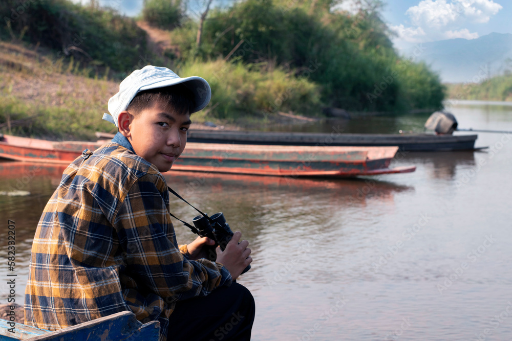 Asian boy in plaid shirt and cap, sitting and holding binoculars on bow of a boat parked beside a river to observe birds flying in the sky and fish swimming in the river during his summer holidays. 