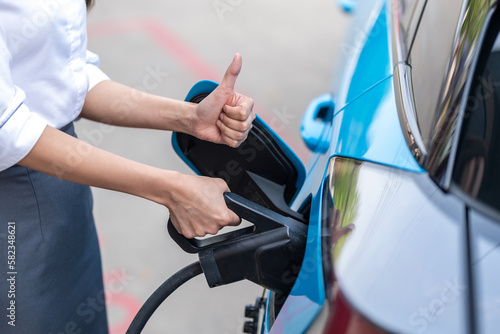 Woman plugging in the charger into a socket of her blue electric car at a charging station in the street © ND STOCK