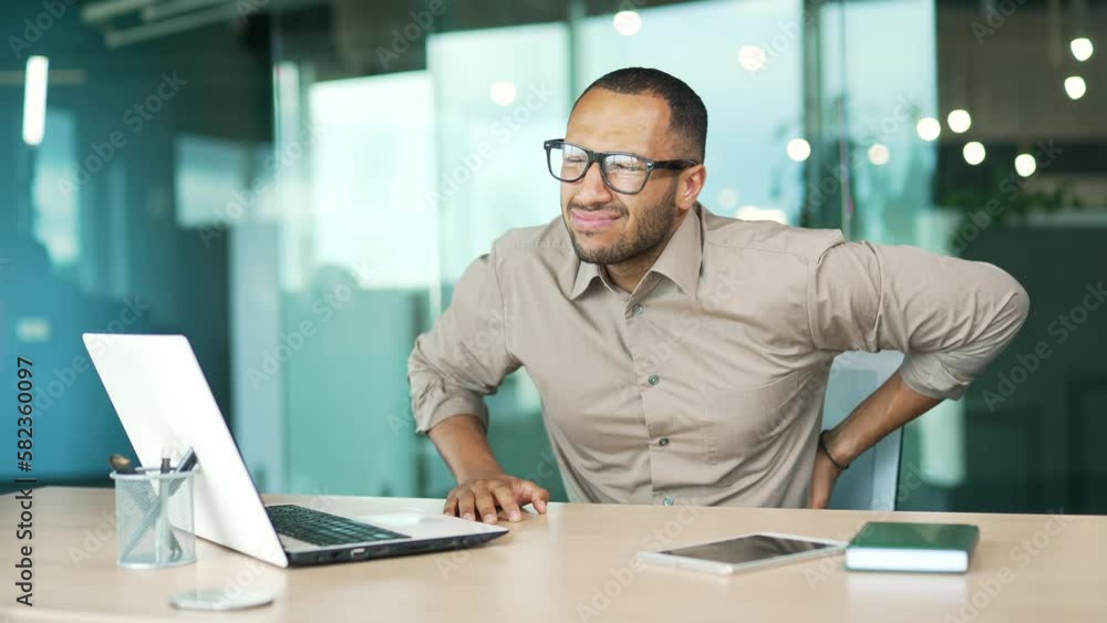 A young adult man has back pain while sitting at a workplace in a modern office. A worker holds his lower back with his hands. He suffers from sciatica, has problems due to a sedentary lifestyle A young adult man has back pain while sitting at a workplace in a modern office. A worker holds his lower back with his hands. He suffers from sciatica, has problems due to a sedentary lifestyle