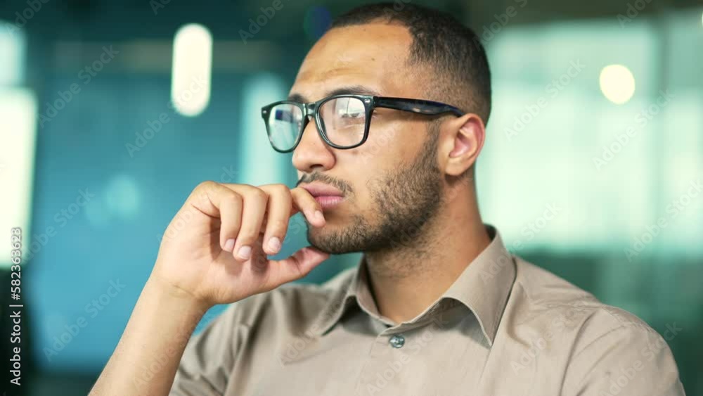 Close up portrait. Young adult man in a shirt and glasses looking seriously to the side while sitting in office. Thoughtful handsome mixed race businessman holding hand near face. Side view. Head shot Close up portrait. Young adult man in a shirt and glasses looking seriously to the side while sitting in office. Thoughtful handsome mixed race businessman holding hand near face. Side view. Head shot