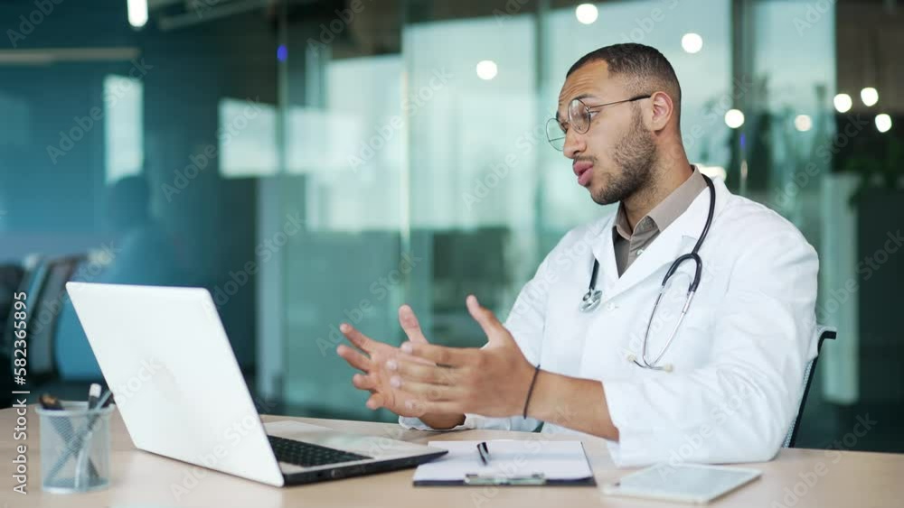 Young smiling doctor is consulting a patient via video call on a laptop while sitting at a workplace in a clinic. Therapist of mixed race in glasses and a white coat speaks, gives advice on treatment Young smiling doctor is consulting a patient via video call on a laptop while sitting at a workplace in a clinic. Therapist of mixed race in glasses and a white coat speaks, gives advice on treatment