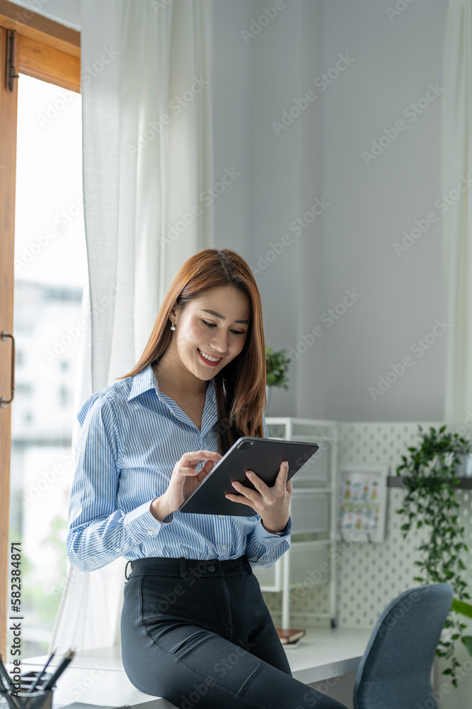 Fototapeta premium A pretty smiling Asian businesswoman holding a tablet to look at financial details, and online delivery orders in the office happily.