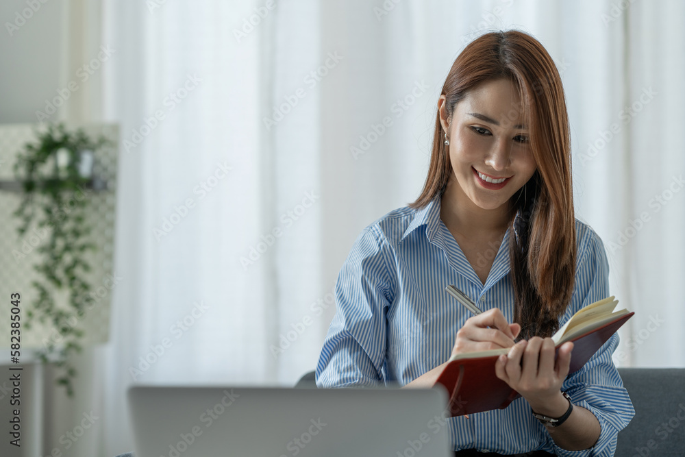 Confident Asian businesswoman sitting and taking notes in financial book Income tax with laptop computer or tablet in a happy office. Confident Asian businesswoman sitting and taking notes in financial book Income tax with laptop computer or tablet in a happy office.