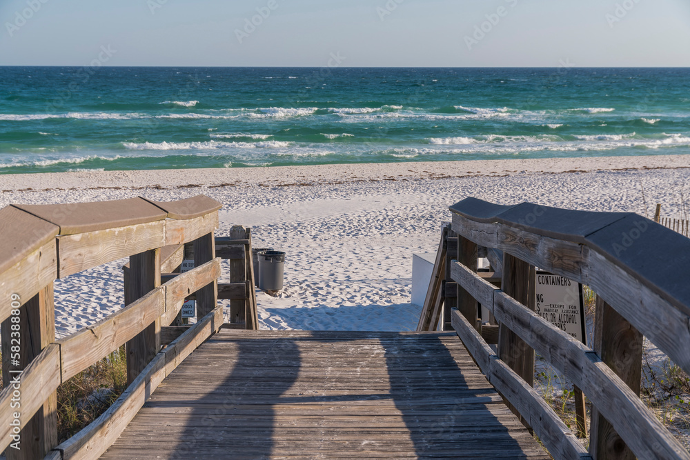 Fototapeta premium Wooden walkway with view of sea and blue sky on a sunny day in Destin Florida. The pathway leads to the sunlit shore and ocean with view of the horizon in the distance.