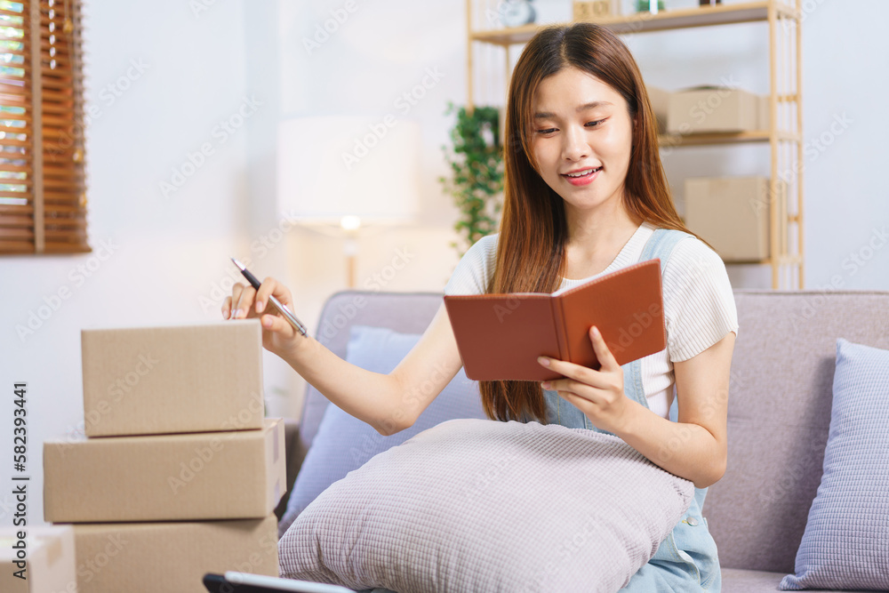 Online seller concept, Female entrepreneur writes on parcel box after reading address on notebook