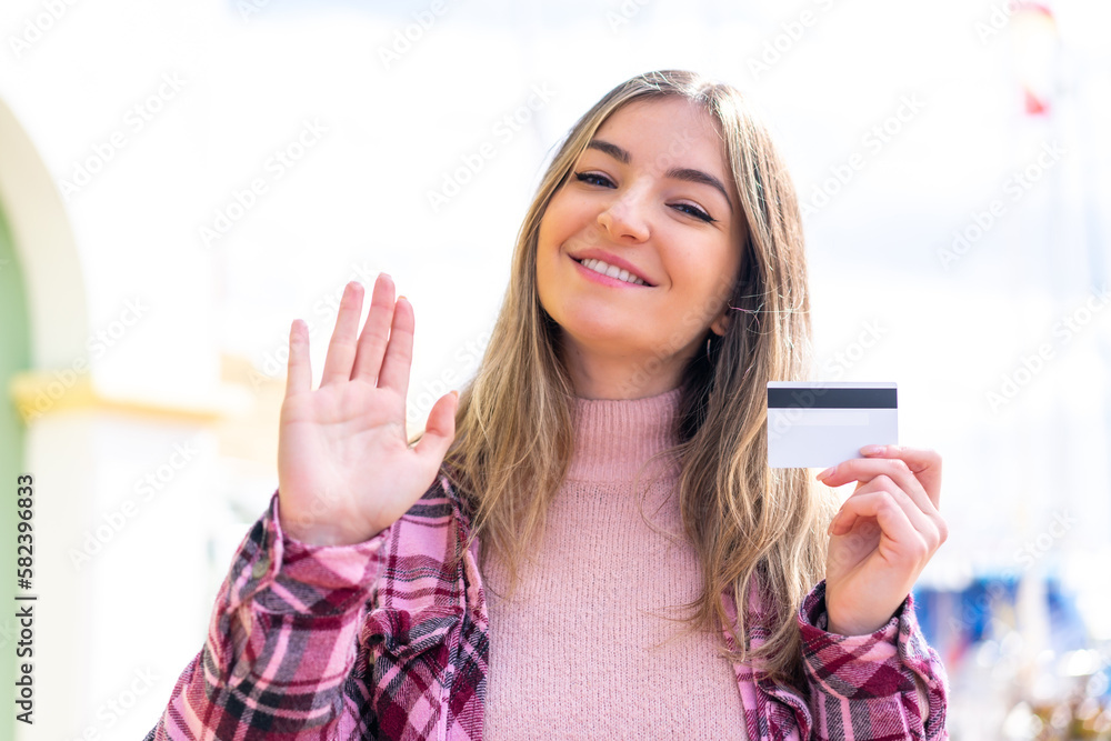 Young pretty Romanian woman holding a credit card at outdoors saluting with hand with happy expression Young pretty Romanian woman holding a credit card at outdoors saluting with hand with happy expression