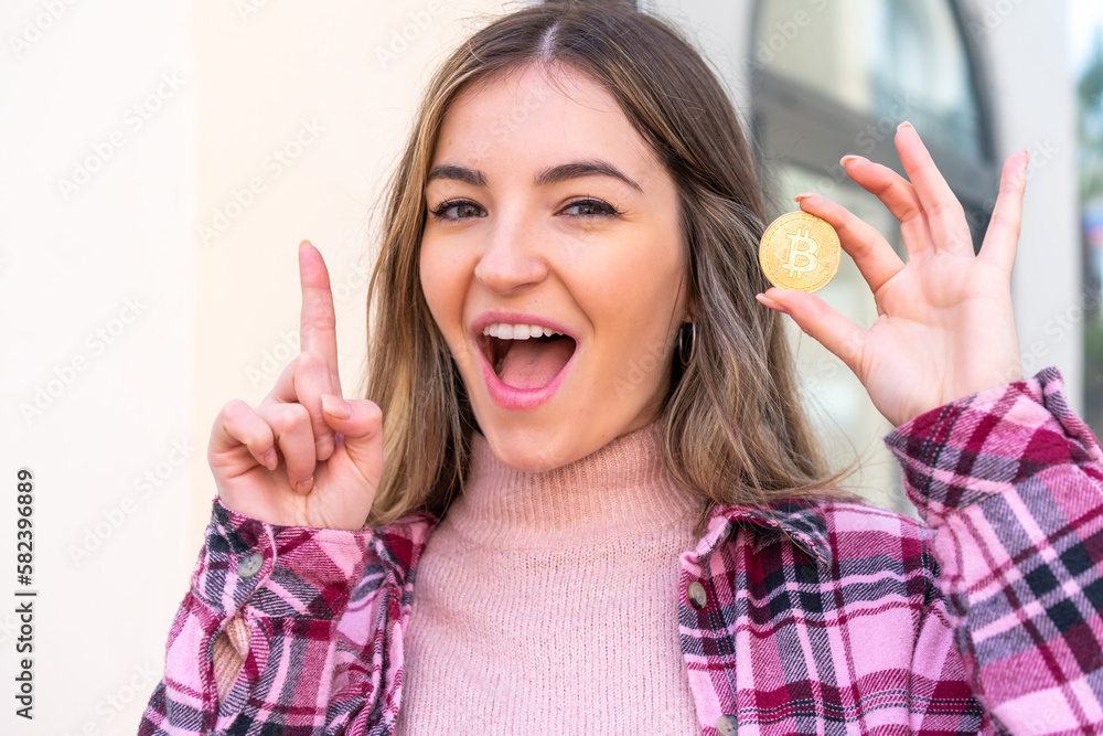 Young pretty Romanian woman holding a Bitcoin at outdoors pointing up a great idea Young pretty Romanian woman holding a Bitcoin at outdoors pointing up a great idea