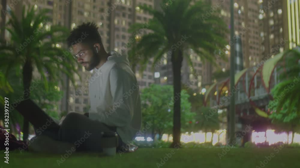 Medium long shot with copy space of male freelancer in hoodie sitting on grass in city park with coffee cup and using the public Internet on laptop while working online in evening Medium long shot with copy space of male freelancer in hoodie sitting on grass in city park with coffee cup and using the public Internet on laptop while working online in evening