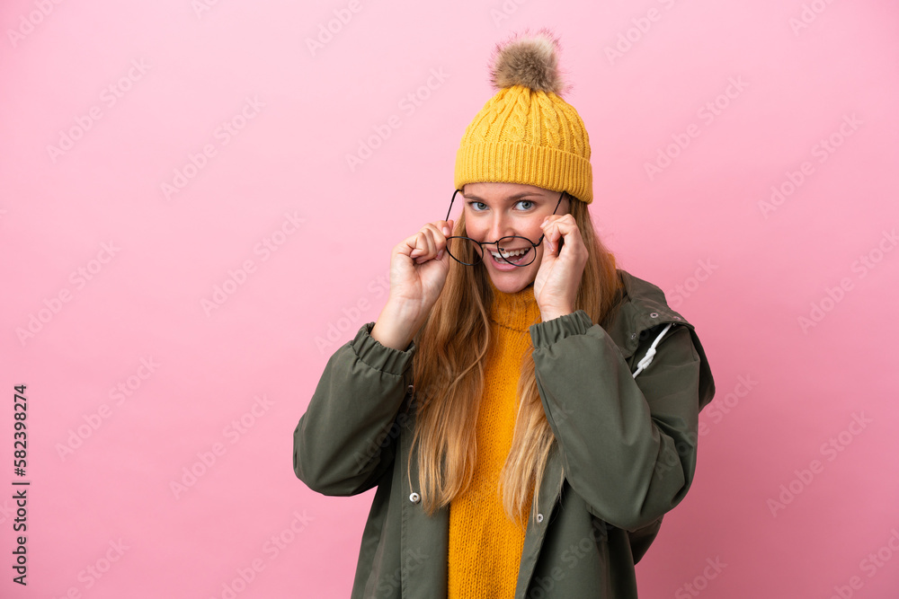 Young blonde woman wearing winter jacket isolated on pink background with glasses and surprised Young blonde woman wearing winter jacket isolated on pink background with glasses and surprised