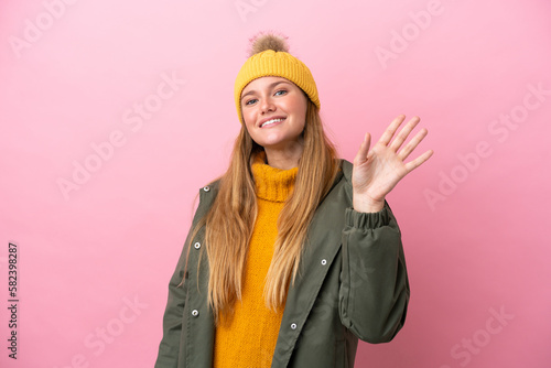 Young blonde woman wearing winter jacket isolated on pink background saluting with hand with happy expression