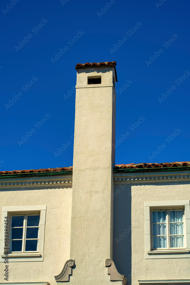 Tall chimney with metal vents and dark adobe roof tiles with visible windows with white frames and beige stucco cement