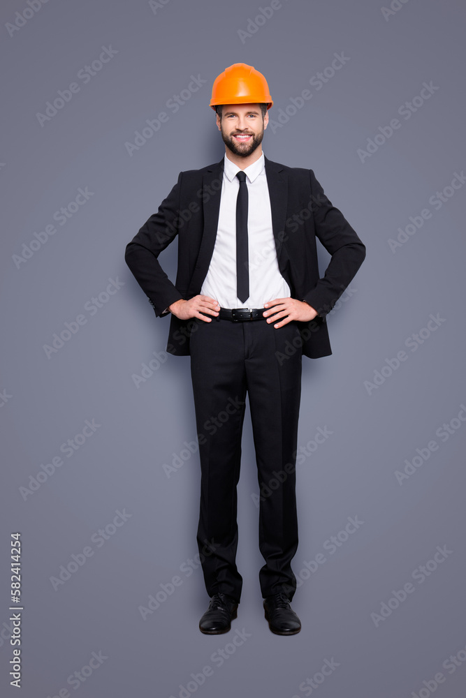 Full length size portrait of attractive cheerful joyful investor in orange hard hat and black tuxedo with tie, looking at camera, isolated on grey background, holding two arms on waist Full length size portrait of attractive cheerful joyful investor in orange hard hat and black tuxedo with tie, looking at camera, isolated on grey background, holding two arms on waist