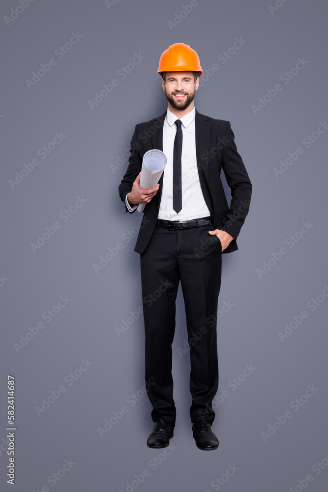 Full length size portrait of attractive cheerful joyful investor in orange hard hat and black tux, tuxedo with tie, looking at camera, isolated on grey background, having paper roll in hand Full length size portrait of attractive cheerful joyful investor in orange hard hat and black tux, tuxedo with tie, looking at camera, isolated on grey background, having paper roll in hand