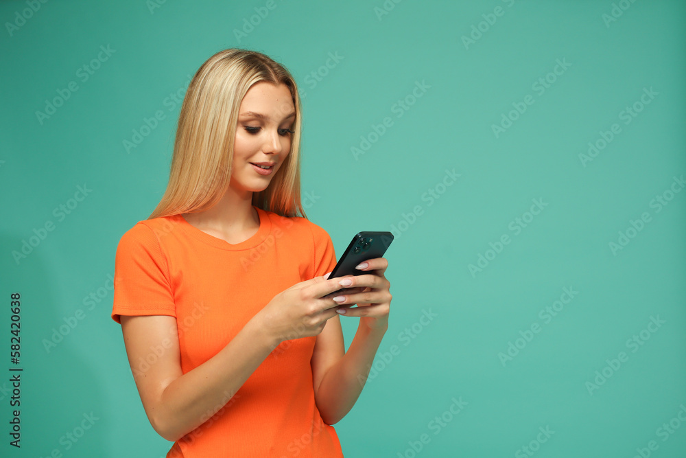 Beautiful blonde girl in an orange t-shirt and jeans with a black phone. Blue background. Studio. Beautiful blonde girl in an orange t-shirt and jeans with a black phone. Blue background. Studio.