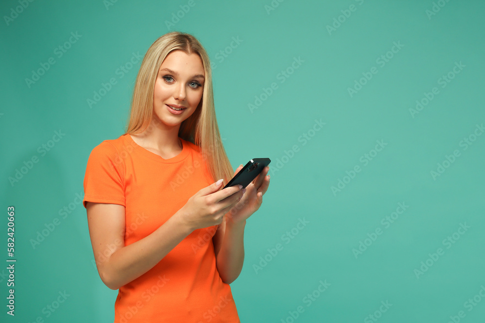 Beautiful blonde girl in an orange t-shirt and jeans with a black phone. Blue background. Studio. Beautiful blonde girl in an orange t-shirt and jeans with a black phone. Blue background. Studio.
