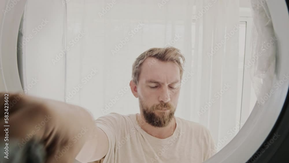 Close-up of young adult bearded man opens door of the dryer machine and takes off his clothes. View from the inside a dryer. Laundry day concept
