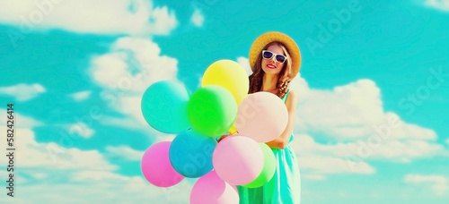 Happy smiling young woman with bunch of colorful balloons wearing a summer straw hat on the field on a blue sky background