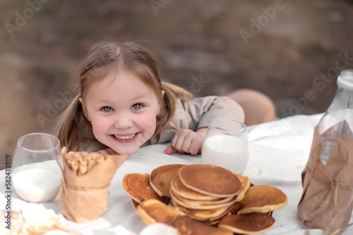 Little cute girl looking at camera and smiling sincerely, sitting at Easter table with pancakes, milk and eggs decorated in rustic style