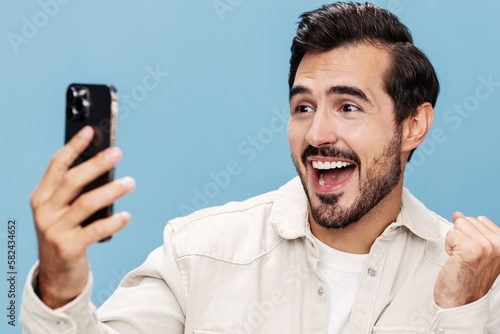 Portrait of a brunette man close-up looking at the phone screen, surprise and open mouth happiness, on a blue background in a white T-shirt and jeans, copy space