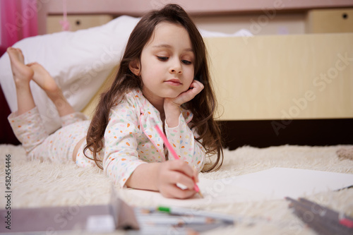 Caucasian little child, a baby girl in stylish pajamas with multicolored dots, drawing picture with colorful pencils indoors in her bedroom. Art. Creativity. Children. Lifestyles. Leisure. Hobby