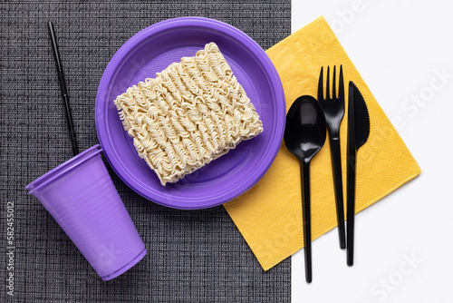 Noodles in a plastic bowl, a lilac cup with a drinking straw, black cutlery on a yellow napkin. Disposable tableware, recycling concept.
