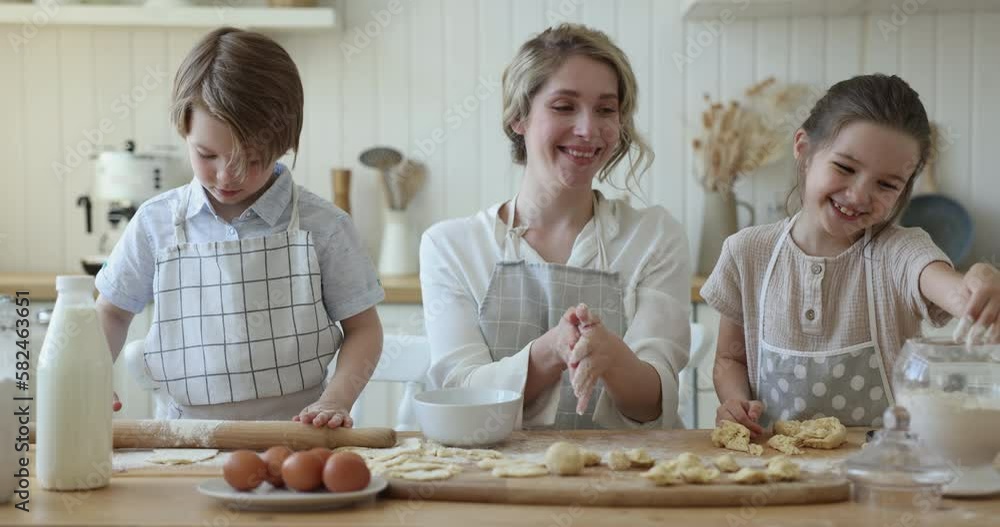 Pretty young mom and little boy and girl baking cookies together, rolling, flouring dough, cooking at kitchen table with floury mess, bakery food ingredients, eggs, milk, talking, smiling Pretty young mom and little boy and girl baking cookies together, rolling, flouring dough, cooking at kitchen table with floury mess, bakery food ingredients, eggs, milk, talking, smiling