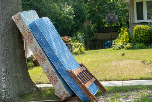 An Old Mattress, box spring and wooden frame by a curb in front of a house near the street waiting to be picked up as trash