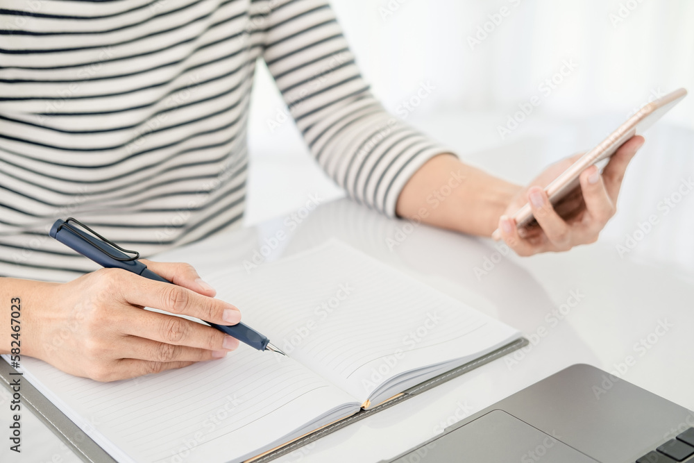 Asian businesswoman in casual clothes writing notes in notepad while working on the phone and laptop , close up clean image in front of the window in soft light , . Asian businesswoman in casual clothes writing notes in notepad while working on the phone and laptop , close up clean image in front of the window in soft light , .