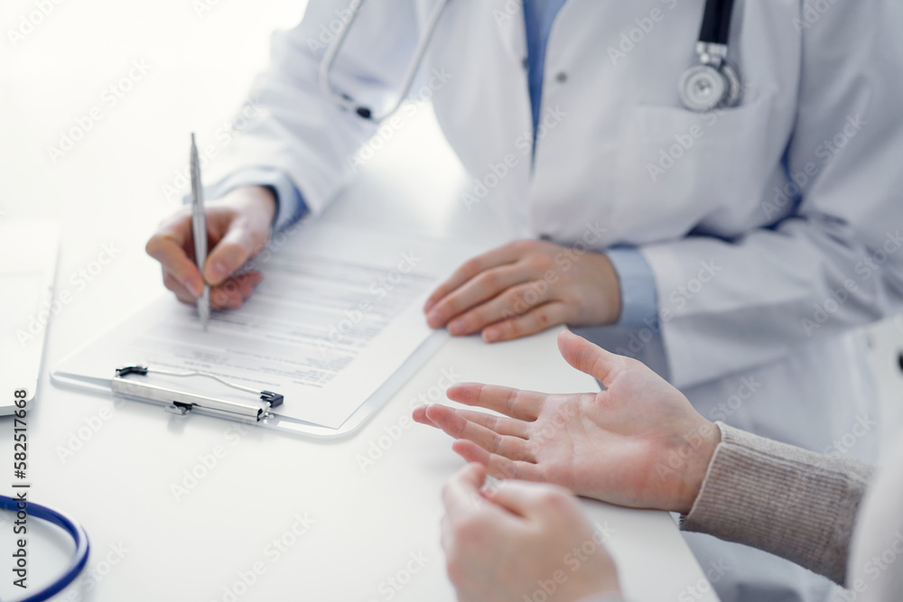 Doctor and patient sitting at the table in clinic. The focus is on female physician's hands filling up the medication history record form or checklist, close up. Medicine concept Doctor and patient sitting at the table in clinic. The focus is on female physician's hands filling up the medication history record form or checklist, close up. Medicine concept