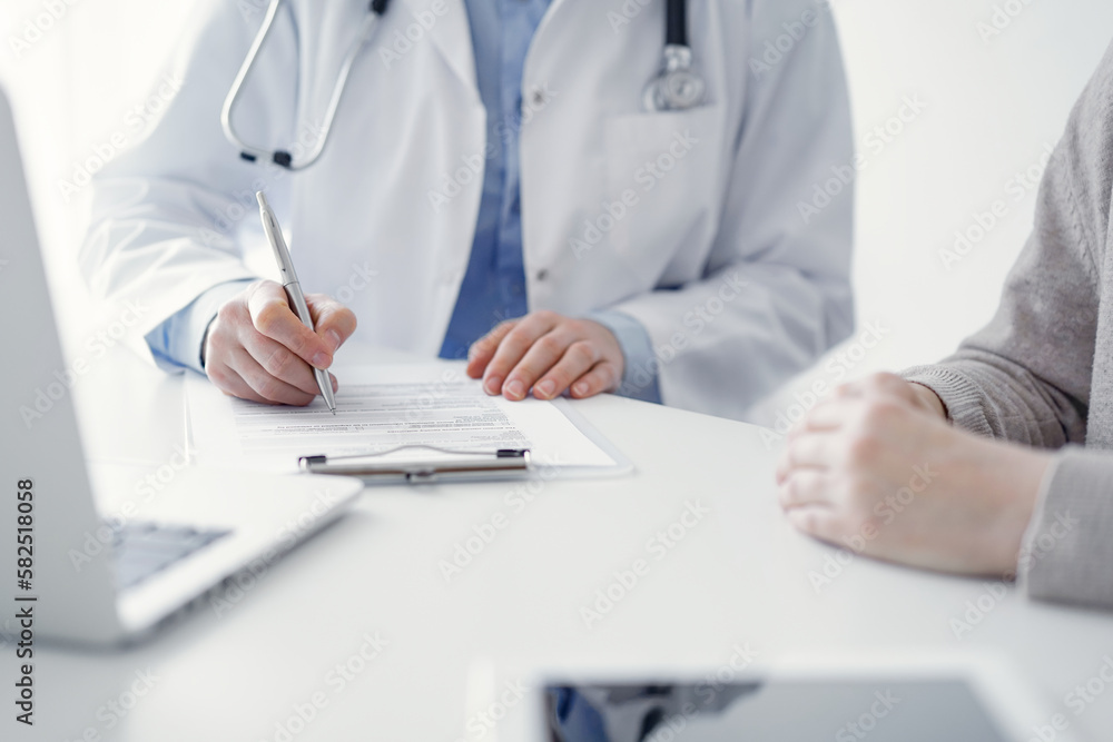 Doctor and patient sitting at the table in clinic. The focus is on female physician's hands filling up the medication history record form or checklist, close up. Medicine concept Doctor and patient sitting at the table in clinic. The focus is on female physician's hands filling up the medication history record form or checklist, close up. Medicine concept