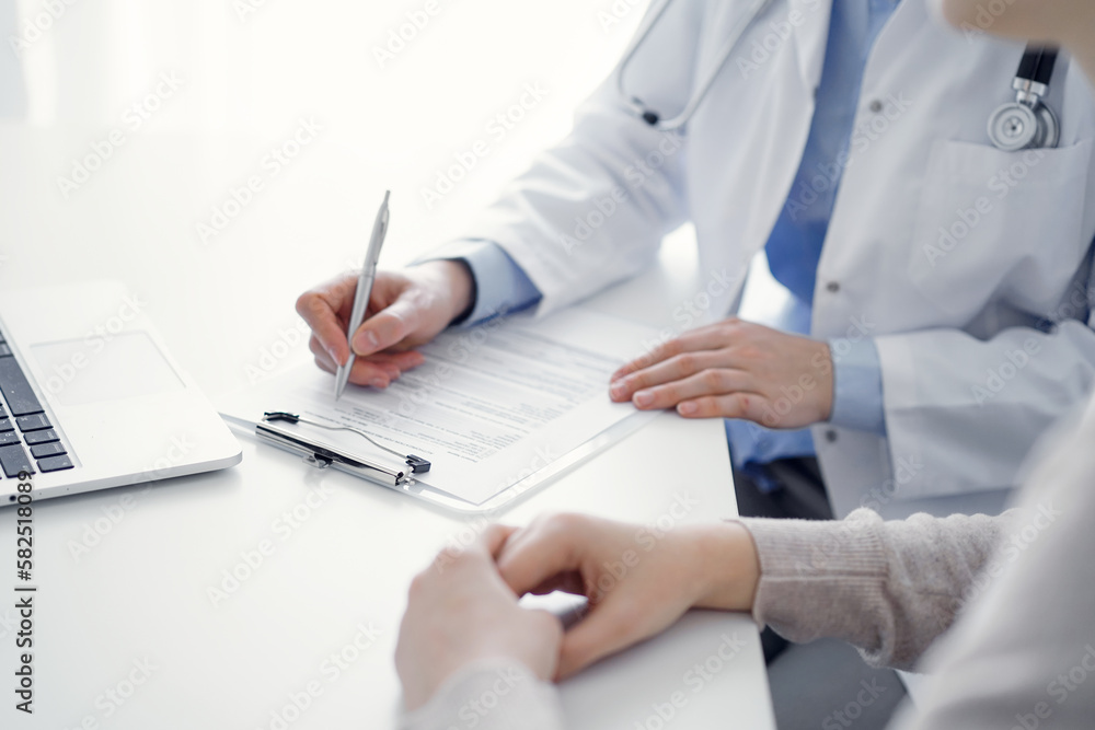 Doctor and patient sitting at the table in clinic. The focus is on female physician's hands filling up the medication history record form or checklist, close up. Medicine concept Doctor and patient sitting at the table in clinic. The focus is on female physician's hands filling up the medication history record form or checklist, close up. Medicine concept