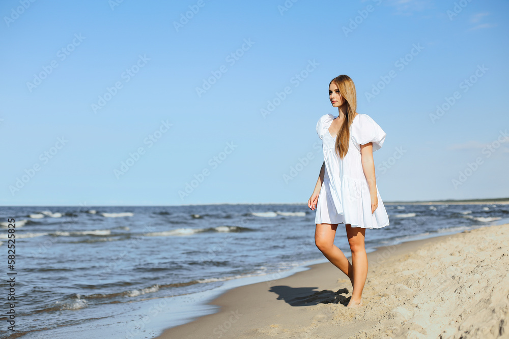 Happy smiling beautiful woman is walking on the ocean beach in a white summer dress