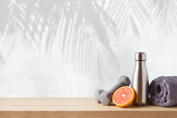Summer fitness background with bottle of water, orange and dumbbells on wooden table over wall with palm tree shadows