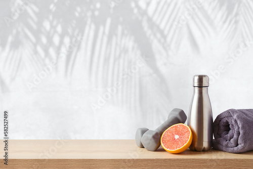 Summer fitness background with bottle of water, orange and dumbbells on wooden table over wall with palm tree shadows
