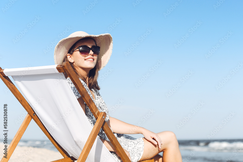 Happy brunette woman wearing sunglasses and hat relaxing on a wooden deck chair at the ocean beach